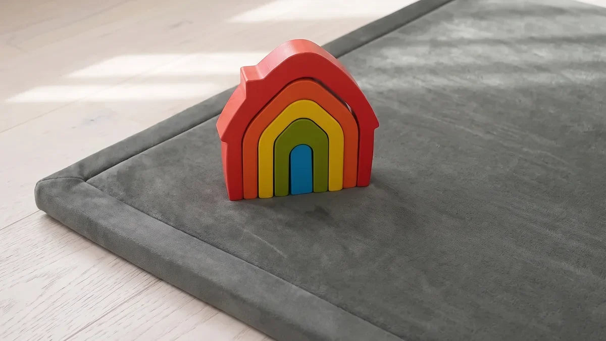 Maison gigogne en bois, cinq maisons rouge, orange, jaune, verte et bleue emboîtées, posée près du coin d'un tapis d'éveil gris en velours