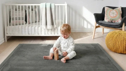 Enfant assis en pyjama blanc qui empile des cubes en bois sur un tapis d'éveil gris, un lit à barreaux blanc et un fauteuil derrière lui