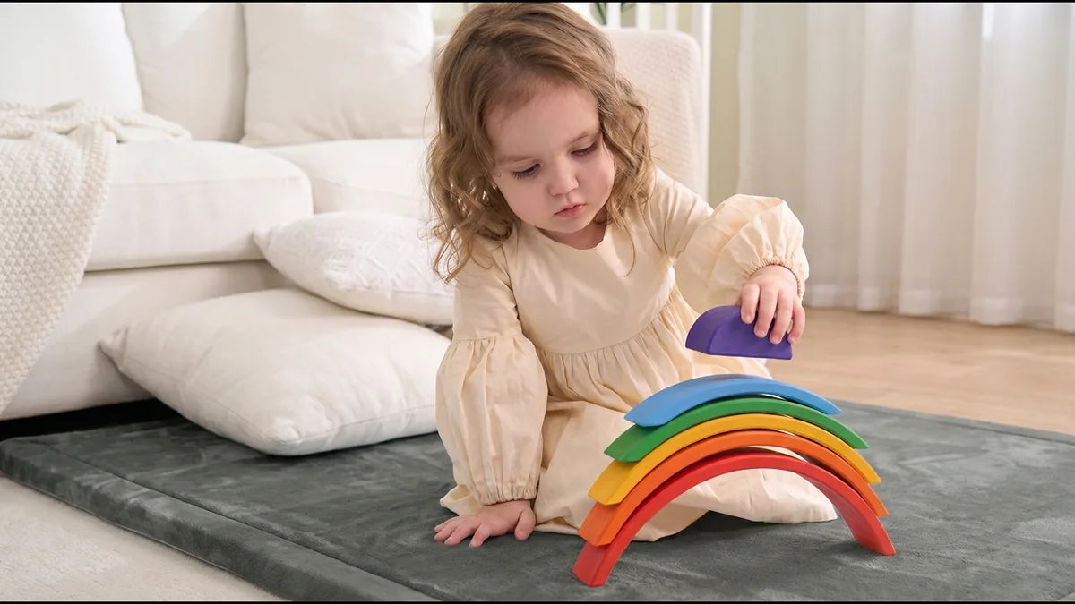 Petite fille assise sur un tapis de jeu gris en velours, qui pose la petite arche violette sur un arc-en-ciel en bois peint