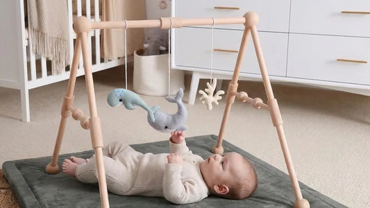 Bébé allongé sur le dos sur un tapis d'éveil gris, sous une arche en bois à perles, la main tendue vers des baleines en feutrine