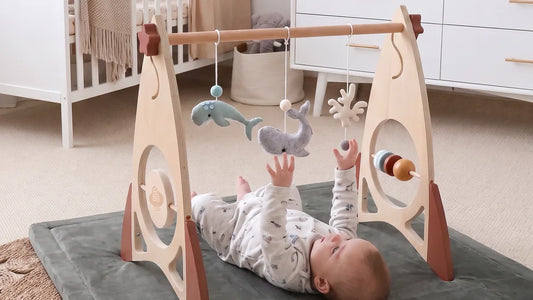Bébé allongé sur le dos sur un tapis d'éveil gris, sous une arche en bois en forme de fusée, les mains vers des baleines en feutrine