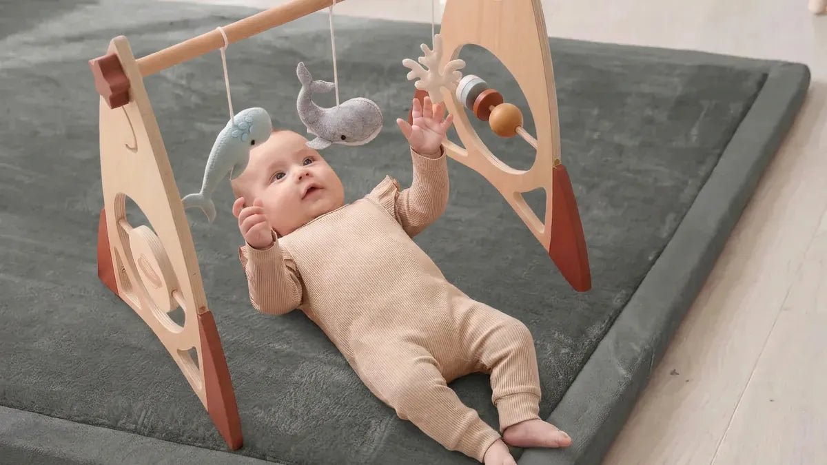 Bébé allongé sur le dos sous une arche d'éveil en bois, la main tendue vers des baleines en feutrine suspendues