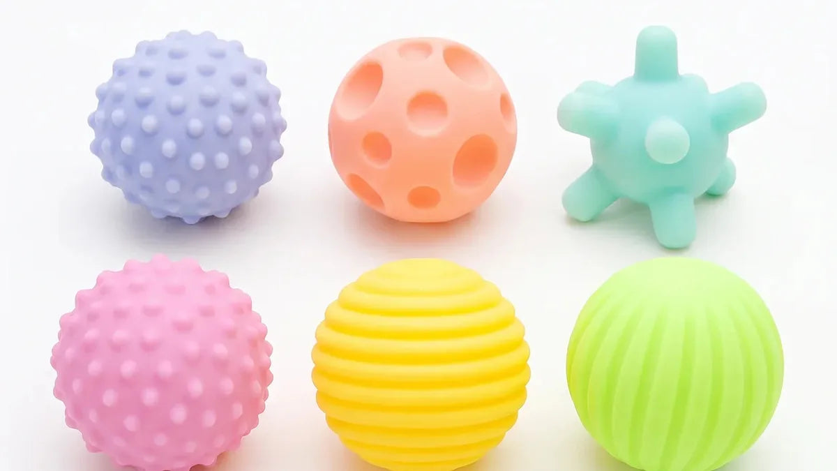 Six balles en silicone pastel sur fond blanc, une à picots lavande, une pêche à creux, une étoile menthe, une rose à picots, une jaune à stries et une verte à côtes