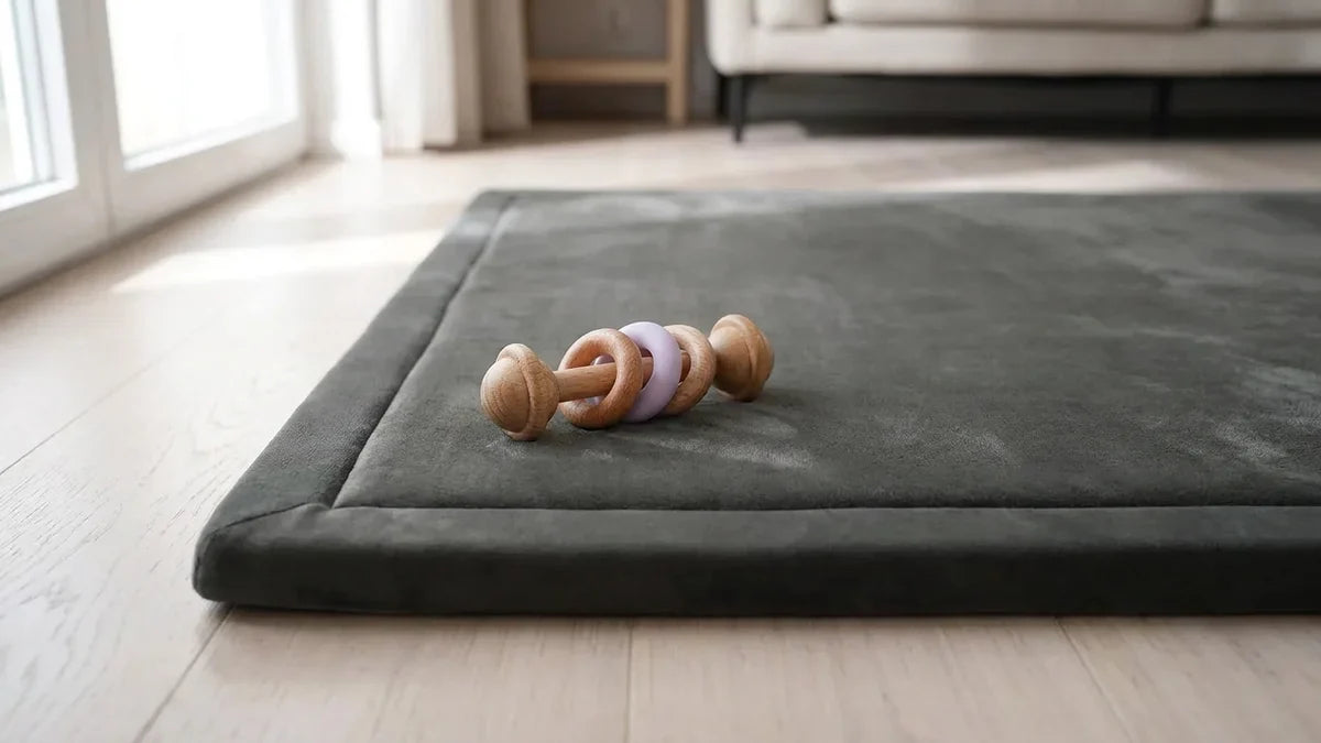 Hochet en bois de hêtre à anneau en silicone violet posé au bord d'un tapis de jeu gris en velours, dans un salon clair