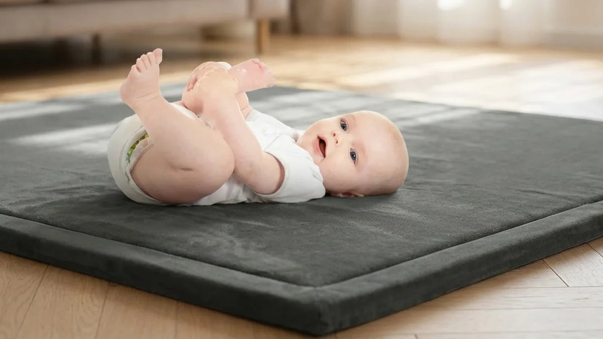 Bébé en body blanc allongé sur le dos sur un tapis d'éveil gris, qui sourit, les jambes levées