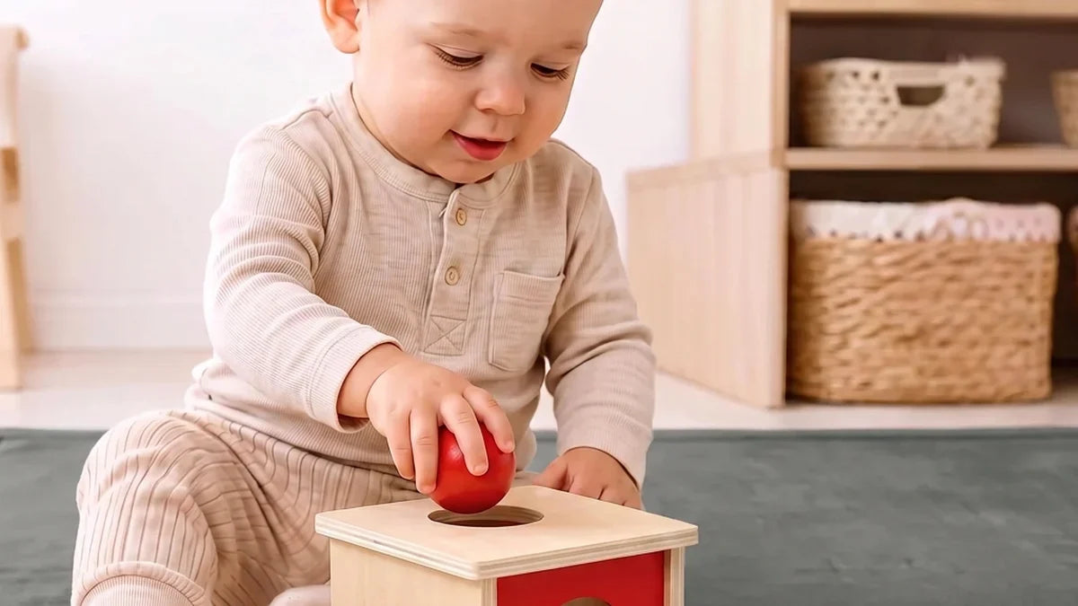 Enfant assis sur un tapis gris en velours qui pousse une balle rouge dans le trou d'une boîte à permanence en bois
