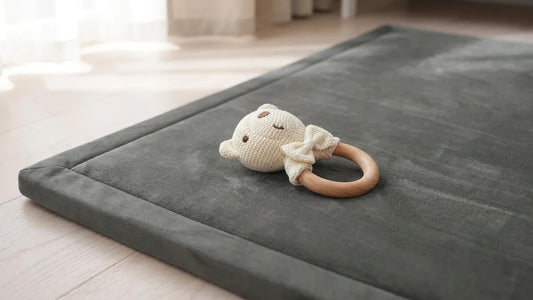 Hochet ours en coton crocheté avec son anneau en bois, posé sur le coin d'un tapis d'éveil en velours gris