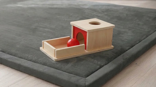 Boîte de permanence en bois clair avec son ouverture en arche et sa balle rouge sur le plateau, posée sur le coin d'un tapis d'éveil gris