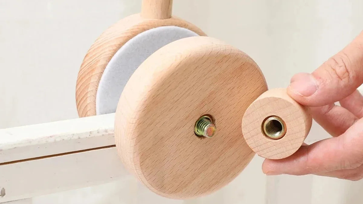 Main qui visse la molette en bois sur la pince ronde feutrée d'un support de mobile, posée sur la barre blanche d'un lit
