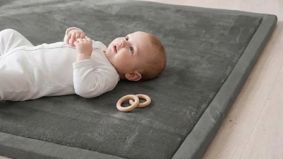 Bébé en grenouillère blanche allongé sur le dos sur un tapis d'éveil gris posé sur un parquet, deux anneaux en bois près de sa main