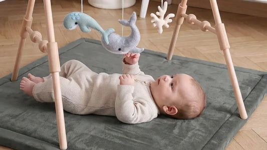 Bébé allongé sur le dos sur un tapis d'éveil gris, sous une arche en bois, la main tendue vers des baleines en feutrine suspendues