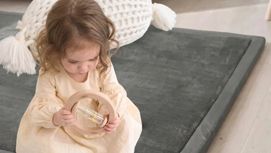 Petite fille assise sur un tapis de jeu gris en velours, un hochet bâton de pluie en bois tenu à deux mains