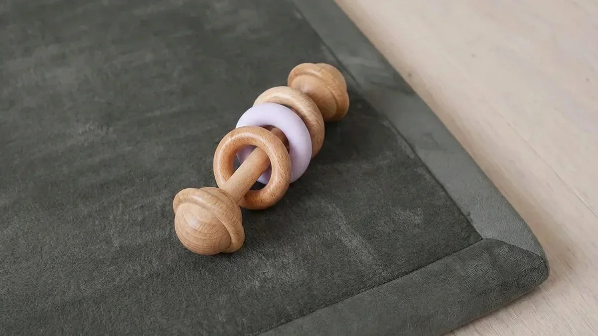 Hochet en hêtre tourné avec deux anneaux en bois et un anneau en silicone lavande, posé sur le bord d'un tapis gris en velours