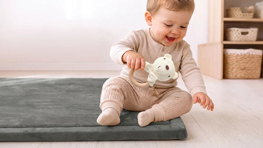Bébé assis sur un tapis d'éveil gris, qui tient un hochet en coton crocheté en forme d'ours avec un anneau en bois