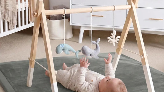 Bébé allongé sur le dos sur un tapis d'éveil gris, sous une arche en bois aux pieds blancs, les mains tendues vers des baleines en feutrine