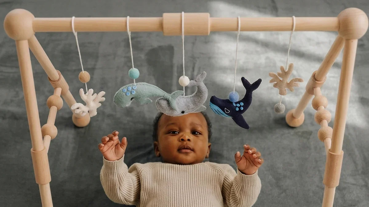 Bébé allongé sur le dos sur un tapis d'éveil gris en velours, les deux mains levées vers des baleines en feutrine suspendues à une arche en bois