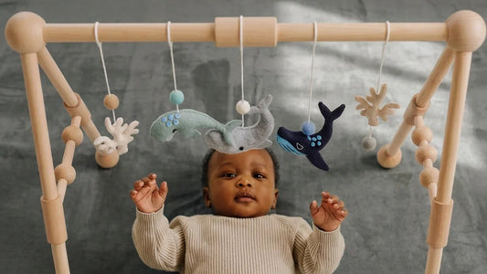 Bébé allongé sur le dos sur un tapis d'éveil gris en velours, les deux mains levées vers des baleines en feutrine suspendues à une arche en bois