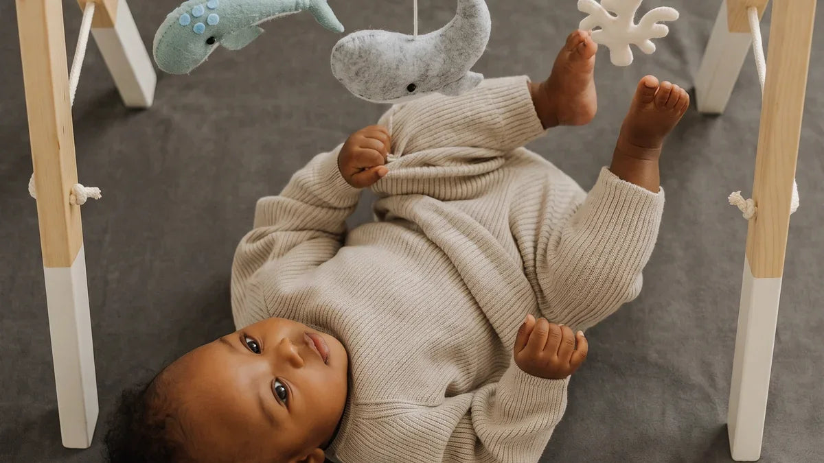 Bébé allongé sur le dos sur un tapis d'éveil gris en velours, les pieds levés vers un corail en feutrine suspendu à une arche en bois aux pieds blancs