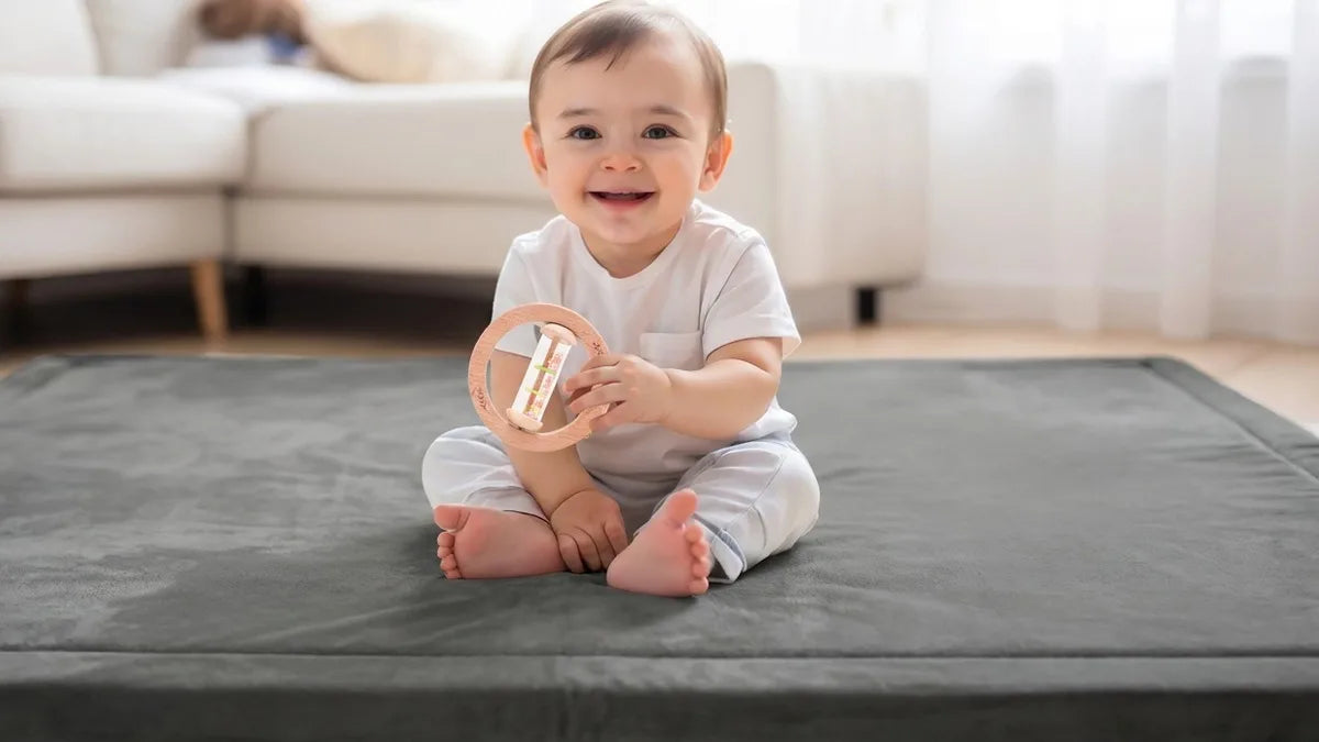 Bébé assis sur un tapis d'éveil gris devant un canapé, qui sourit en tenant un hochet bâton de pluie en bois à deux mains