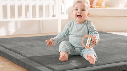 Bébé assis seul sur un tapis d'éveil gris en velours, souriant, un hochet bâton de pluie en bois dans la main