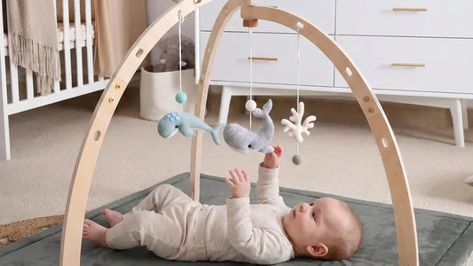 Bébé allongé sur le dos sur un tapis d'éveil gris, sous une arche en bois, la main tendue vers des baleines en feutrine suspendues