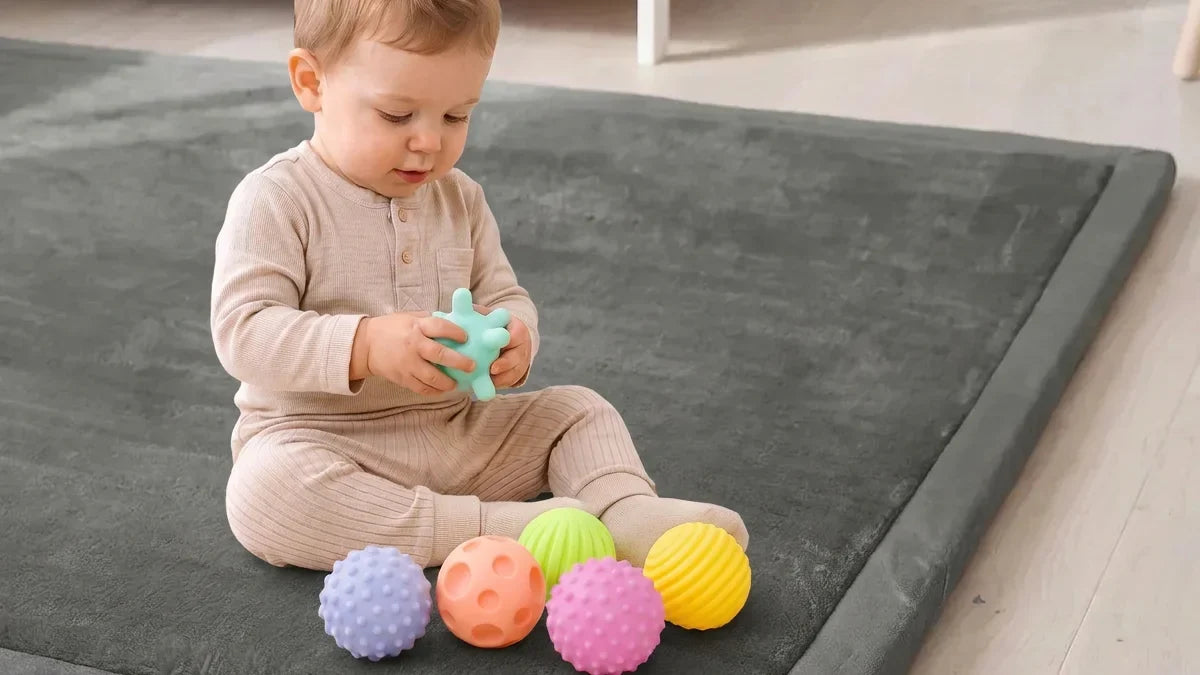 Bébé assis sur un tapis d'éveil gris, une balle en silicone à picots dans les mains, cinq autres balles devant lui