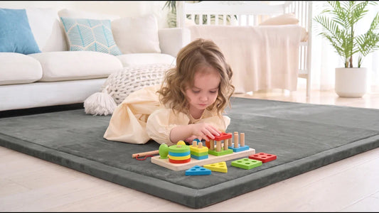 Petite fille allongée sur un tapis de jeu gris en velours, qui enfile une forme rouge sur une planche en bois à colonnes, dans un salon clair