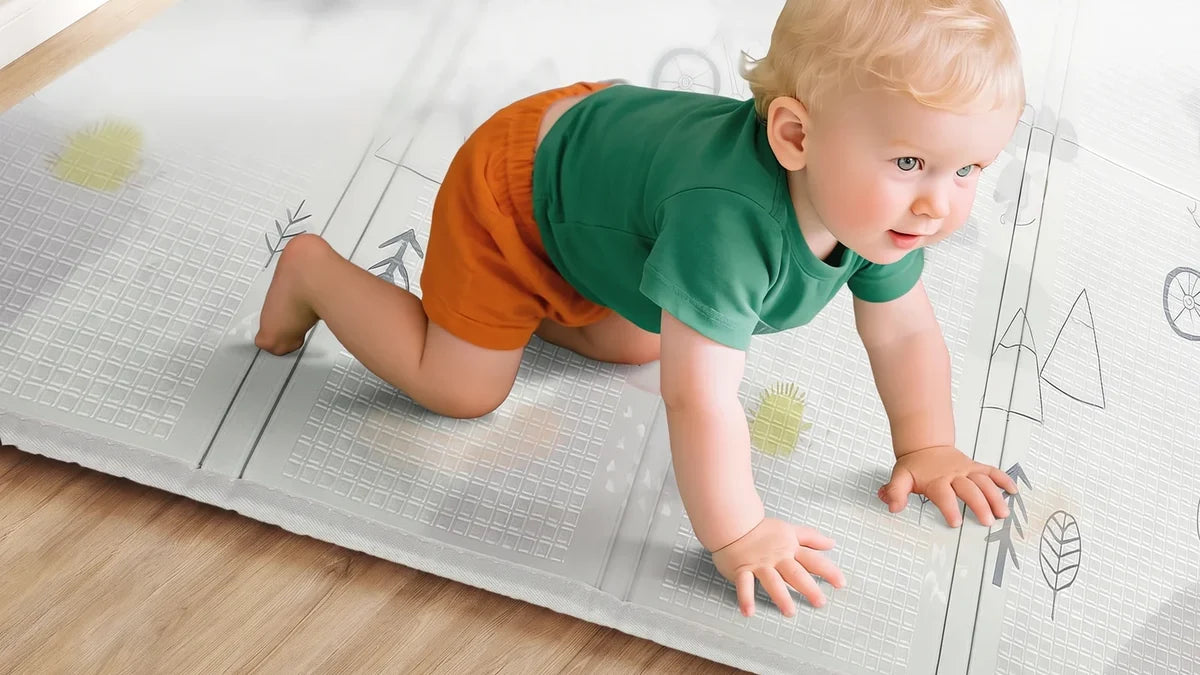 Un bébé en tee-shirt vert avance à quatre pattes sur un tapis d'éveil pliable gris clair imprimé de sapins, posé sur un parquet