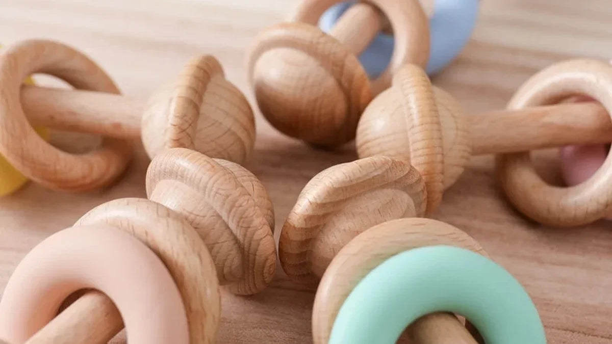 Plusieurs hochets en hêtre posés sur une table en bois clair, chacun avec deux anneaux en bois et un anneau en silicone rose, vert d'eau ou bleu