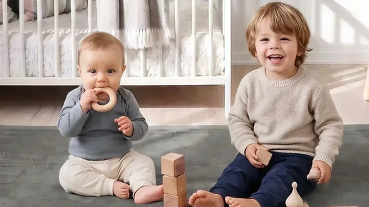 Un bébé assis sur un tapis d'éveil gris porte un anneau en bois à la bouche, à côté d'un enfant plus grand qui tient un cube en bois