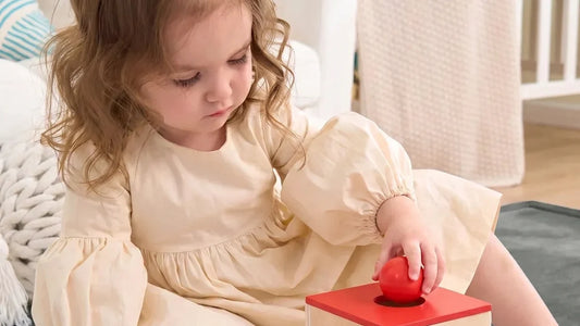 Une petite fille assise sur un tapis d'éveil gris pose une balle rouge dans le trou d'une boîte en bois