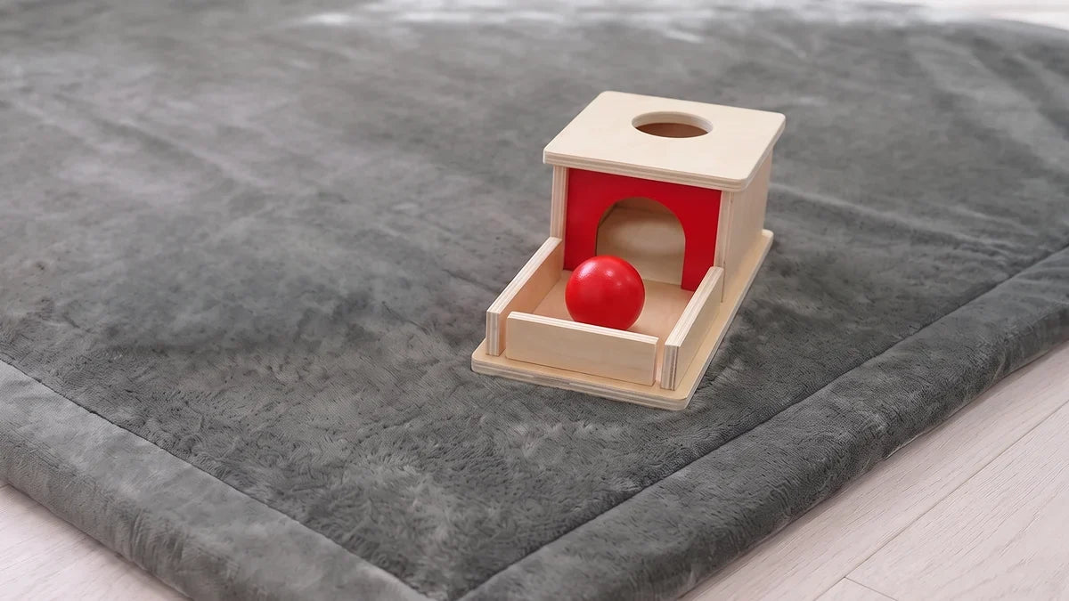 Une boîte de permanence en bois avec sa balle rouge dans le plateau, posée près du coin d'un tapis d'éveil gris
