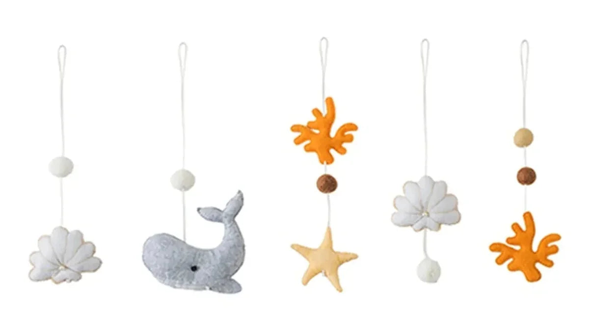 Cinq jouets en feutrine gris, blanc et orange, une baleine, une étoile, des coraux et des coquillages, chacun sur un cordon blanc