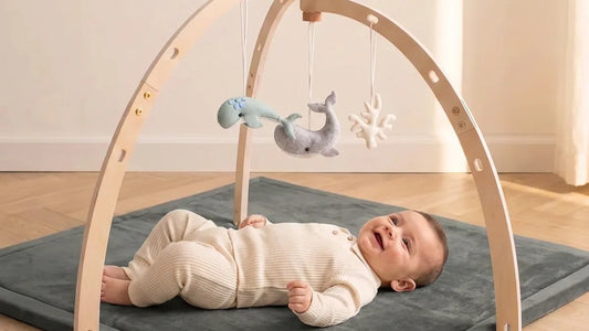 Bébé allongé sur le dos sur un tapis d'éveil gris en velours, sous une arche en bois double arc où pendent des baleines en feutrine