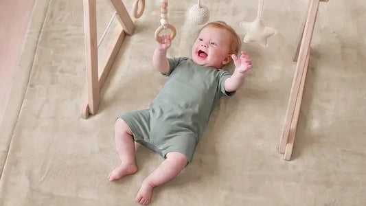 Bébé allongé sur le dos sur un tapis d'éveil en velours beige sable, qui attrape un anneau en bois suspendu à une arche