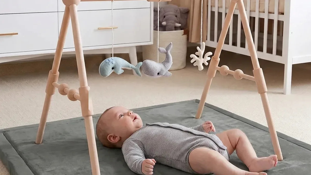 Bébé allongé sur le dos sur un tapis d'éveil gris, sous une arche en bois à perles avec des baleines en feutrine, dans une chambre avec commode et lit à barreaux