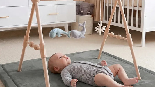 Bébé allongé sur le dos sur un tapis d'éveil gris, sous une arche en bois à perles avec des baleines en feutrine, dans une chambre avec commode et lit à barreaux
