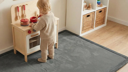 Enfant en pull beige debout sur un tapis d'éveil en velours gris devant une cuisine en bois, dans une chambre au parquet clair