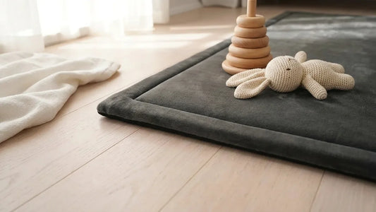 Bord d'un tapis d'éveil en velours gris anthracite sur un parquet clair, une pyramide d'anneaux en bois et un lapin en crochet posés dessus