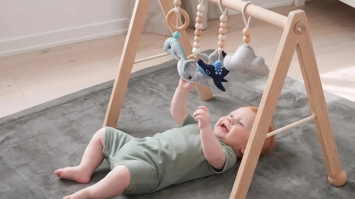 Bébé allongé sur le dos sous une arche d'éveil en bois, sur un tapis d'éveil gris, les mains vers une baleine en feutrine