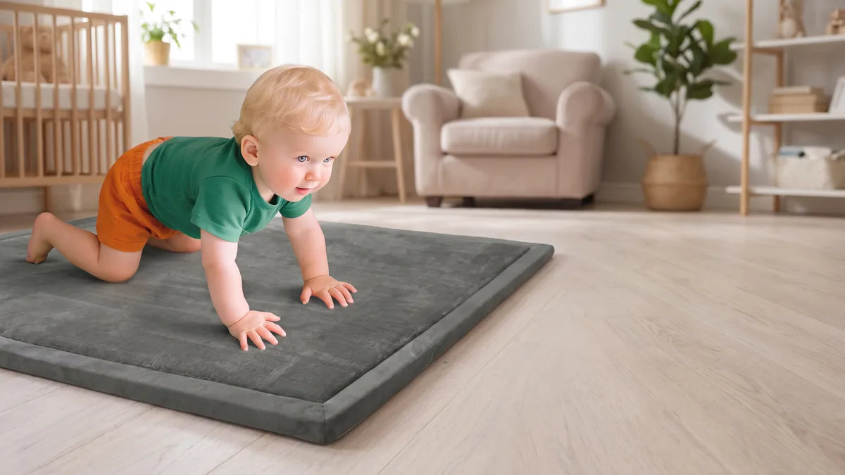 Bébé en t-shirt vert à quatre pattes sur un tapis d'éveil gris en velours posé sur un parquet clair, un fauteuil et un lit à barreaux derrière lui
