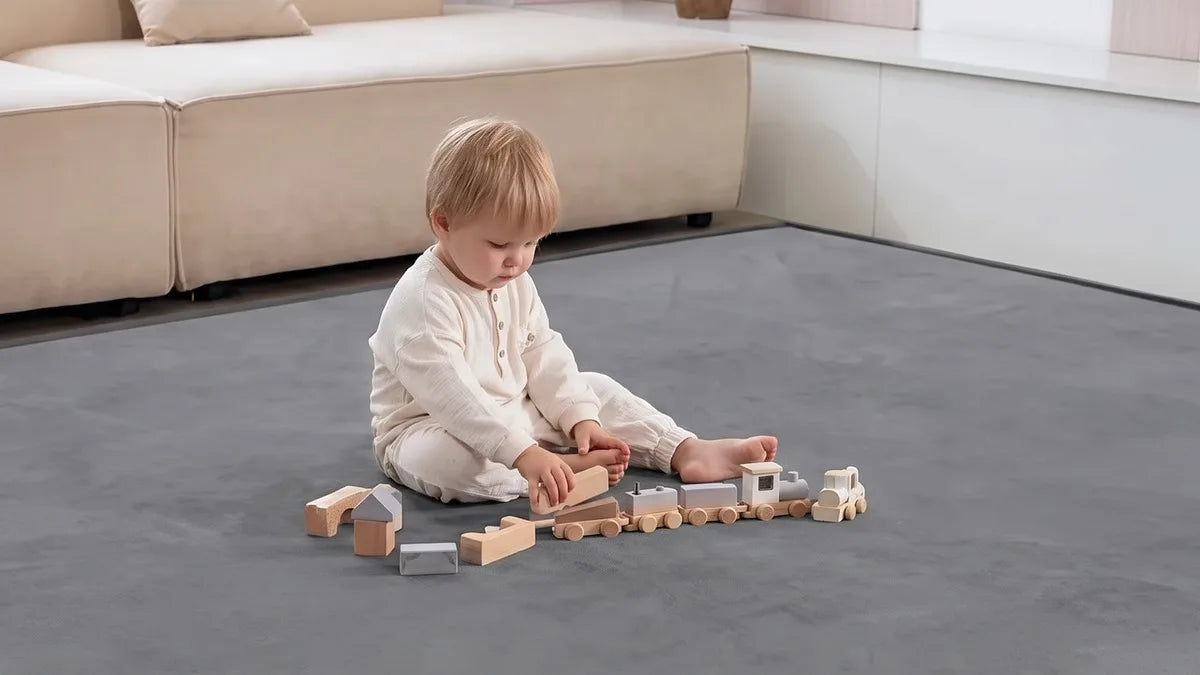 Enfant assis seul sur un tapis d'éveil gris au salon, des blocs et un petit train en bois devant lui