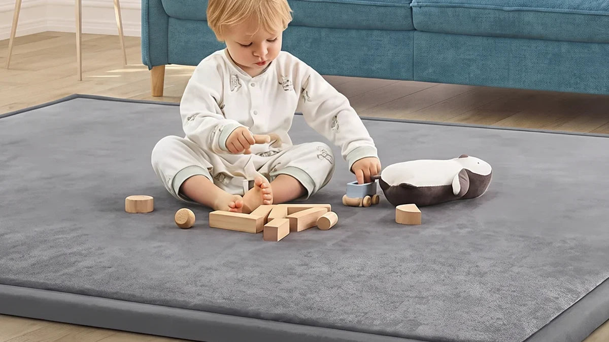Enfant assis sur le bord d'un tapis d'éveil en velours gris anthracite dans un salon, des cubes en bois et une peluche devant lui