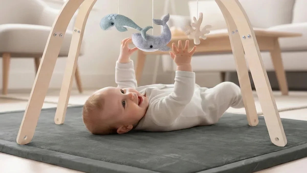Bébé allongé sur le dos sur un tapis d'éveil gris, sous une arche en bois, tend les mains vers deux baleines en feutrine