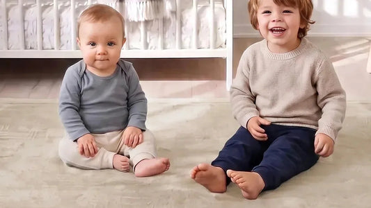 Deux enfants assis pieds nus côte à côte sur un tapis d'éveil beige sable, un lit à barreaux blanc derrière eux