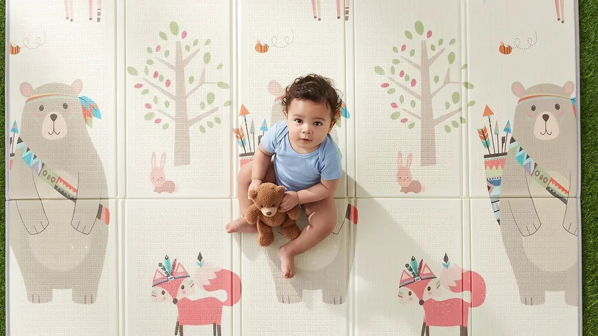 Bébé en body bleu assis avec un ourson en peluche sur un tapis d'éveil pliable imprimé d'ours et de renards, posé sur l'herbe