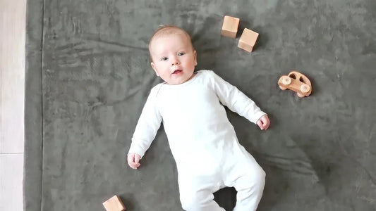 Bébé en grenouillère blanche allongé sur le dos sur un tapis d'éveil gris, cubes et petite voiture en bois autour