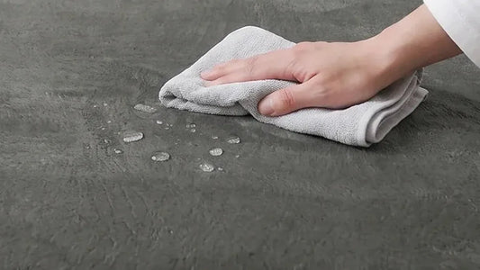 Main qui passe un chiffon humide sur le velours gris d'un tapis d'éveil, gouttes d'eau posées à la surface