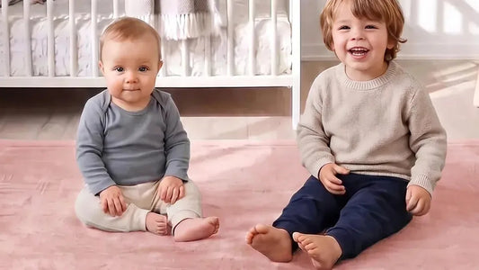 Deux enfants assis côte à côte sur un tapis d'éveil rose poudré, un lit à barreaux blanc derrière eux