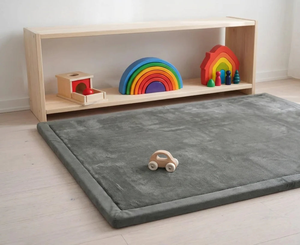 Coin de jeu Montessori, le tapis d'éveil gris au sol devant une étagère basse en bois avec trois jouets
